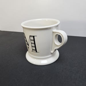 May include: White ceramic mug with a black letter "M" design. The mug has a wide base and a curved handle. The letter "M" is decorated with a black and white striped pattern. The mug is on a dark surface.