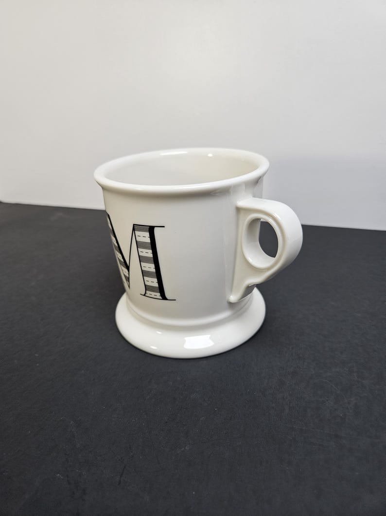May include: White ceramic mug with a black letter "M" design. The mug has a wide base and a curved handle. The letter "M" is decorated with a black and white striped pattern. The mug is on a dark surface.