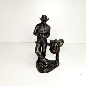 May include: A bronze figurine of a cowboy wearing a hat and leaning against a fence post with a saddle hanging on it.