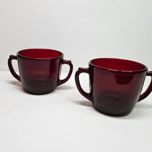May include: Two red glass cups with handles. The cups are made of a dark red glass and have a rounded shape.