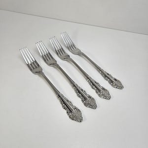 May include: A set of four silver-toned metal forks arranged in a diagonal row. Each fork features a polished finish with intricate, embossed floral and scrollwork detailing along the handle, ending in a rounded, decorative base.