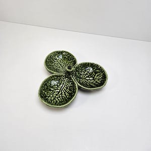 May include: A green ceramic serving dish with three leaf-shaped compartments. The dish has a glossy finish and detailed leaf vein patterns. The three sections are connected at a central point.