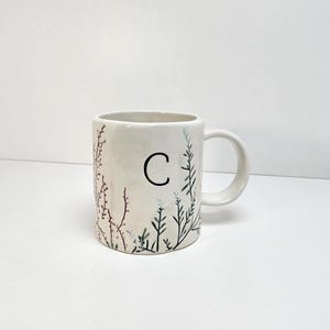 K&ouml;nnte beinhalten: Cremefarbene Keramiktasse mit dem schwarzen Buchstaben "C" und botanischen Illustrationen in Gr&uuml;n und Burgunderrot. Die Tasse hat einen Henkel und ist f&uuml;r Hei&szlig;getr&auml;nke geeignet.