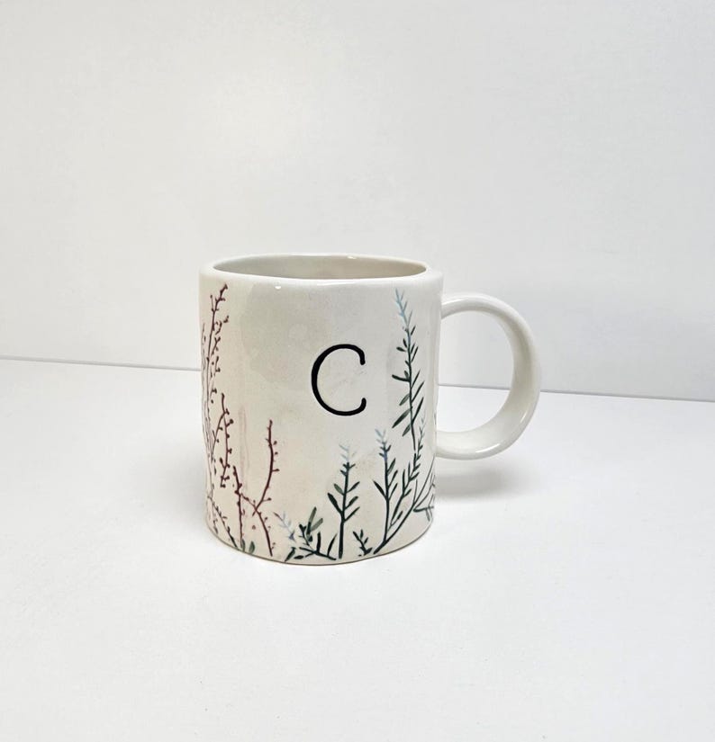 K&ouml;nnte beinhalten: Cremefarbene Keramiktasse mit dem schwarzen Buchstaben "C" und botanischen Illustrationen in Gr&uuml;n und Burgunderrot. Die Tasse hat einen Henkel und ist f&uuml;r Hei&szlig;getr&auml;nke geeignet.