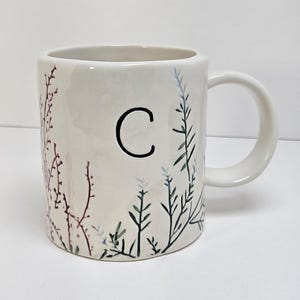 K&ouml;nnte beinhalten: Eine wei&szlig;e Keramiktasse mit Henkel, mit einem schwarzen Buchstaben "C" und bemalten botanischen Designs in Gr&uuml;n-, Blau- und Burgundert&ouml;nen. Die Tasse hat eine glatte, gl&auml;nzende Oberfl&auml;che und ist f&uuml;r hei&szlig;e oder kalte Getr&auml;nke geeignet.