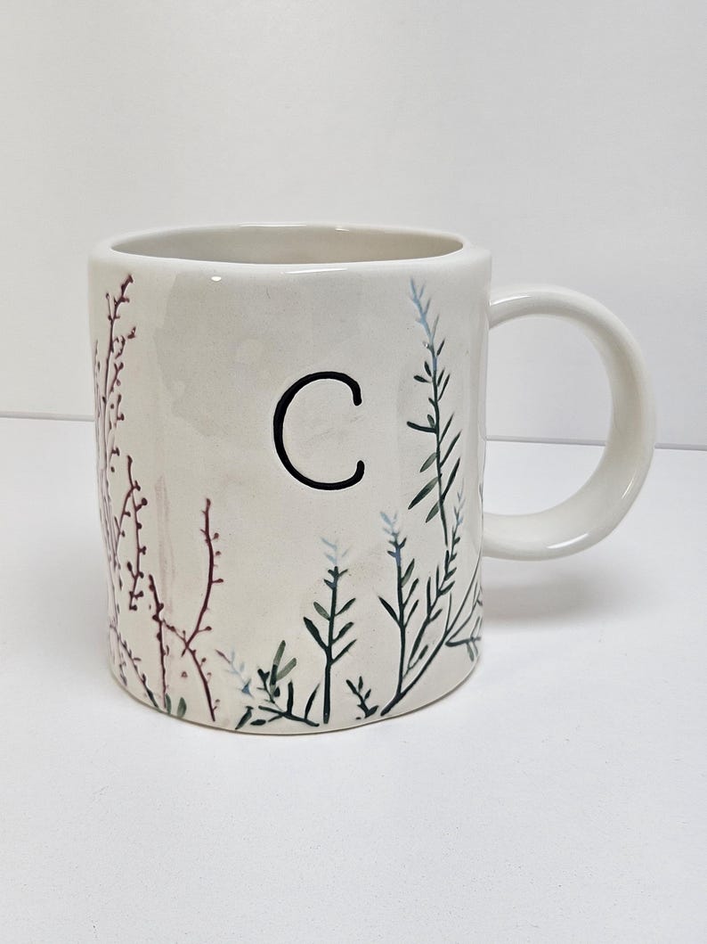 K&ouml;nnte beinhalten: Eine wei&szlig;e Keramiktasse mit Henkel, mit einem schwarzen Buchstaben "C" und bemalten botanischen Designs in Gr&uuml;n-, Blau- und Burgundert&ouml;nen. Die Tasse hat eine glatte, gl&auml;nzende Oberfl&auml;che und ist f&uuml;r hei&szlig;e oder kalte Getr&auml;nke geeignet.