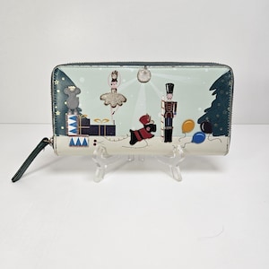 May include: A rectangular wallet with a zipper closure. The wallet features a festive design with a ballerina, nutcracker, and other holiday-themed illustrations. The color palette includes teal, white, and gold accents. The wallet is displayed on a clear stand.