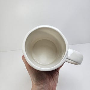 K&ouml;nnte beinhalten: Ein wei&szlig;er Keramikbecher mit gro&szlig;em Henkel. Der Becher wird vor einem wei&szlig;en Hintergrund gehalten. Auch das Innere des Bechers ist wei&szlig;.