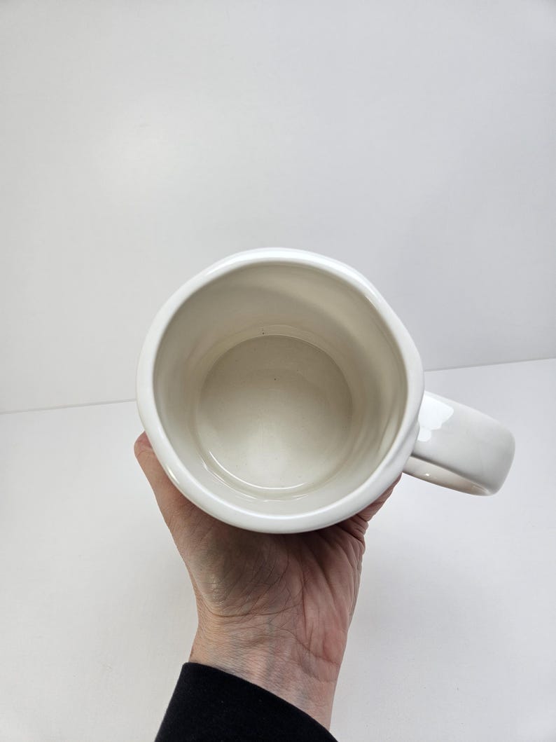 K&ouml;nnte beinhalten: Ein wei&szlig;er Keramikbecher mit gro&szlig;em Henkel. Der Becher wird vor einem wei&szlig;en Hintergrund gehalten. Auch das Innere des Bechers ist wei&szlig;.