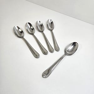 May include: A set of five silver-toned stainless steel spoons arranged on a surface. Each spoon features a polished oval bowl and a handle decorated with a beaded border and a small shell-like motif at the very end of the handle base.