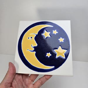 May include: Square ceramic tile featuring a smiling yellow crescent moon and five yellow stars against a dark blue background. The moon and stars are outlined in a darker blue, with a white border.