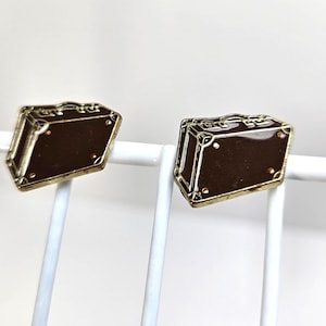 May include: Two gold-toned suitcase-shaped stud earrings. The earrings have a dark brown enamel center and a gold border. The suitcase design includes a handle and corner details. The earrings are displayed on a white wire rack.