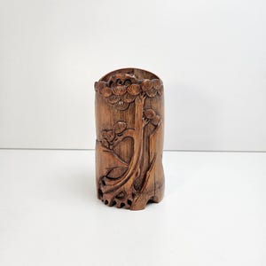 May include: A cylindrical, wooden pen holder with a detailed carved tree design. The pen holder is a warm brown color and features intricate carvings of branches and leaves. The top edge has a scalloped design.
