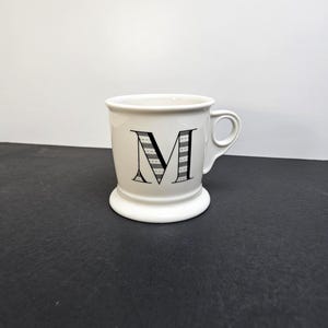May include: A white ceramic mug with a black handle and the text "But First, Coffee" in a black, bold font. The mug is sitting on a wooden surface. The mug is approximately 4 inches tall.
