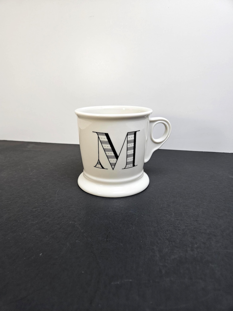 May include: A white ceramic mug with a black handle and the text "But First, Coffee" in a black, bold font. The mug is sitting on a wooden surface. The mug is approximately 4 inches tall.