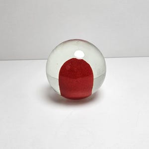 May include: A clear glass paperweight with a vibrant red interior. The red portion is a textured, rounded shape, encased within the transparent glass sphere. The paperweight sits on a white surface.