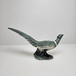 May include: A ceramic figurine of a green pheasant with a long tail. The pheasant is standing on a base with green leaves.