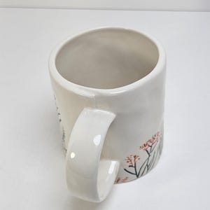 K&ouml;nnte beinhalten: Wei&szlig;er Keramikbecher mit gebogenem Henkel. Der Becher hat ein Blumendesign mit roten und gr&uuml;nen Akzenten. Der Becher ist zylindrisch und hat eine glatte, gl&auml;nzende Oberfl&auml;che. Der Griff ist dick und leicht zu greifen.