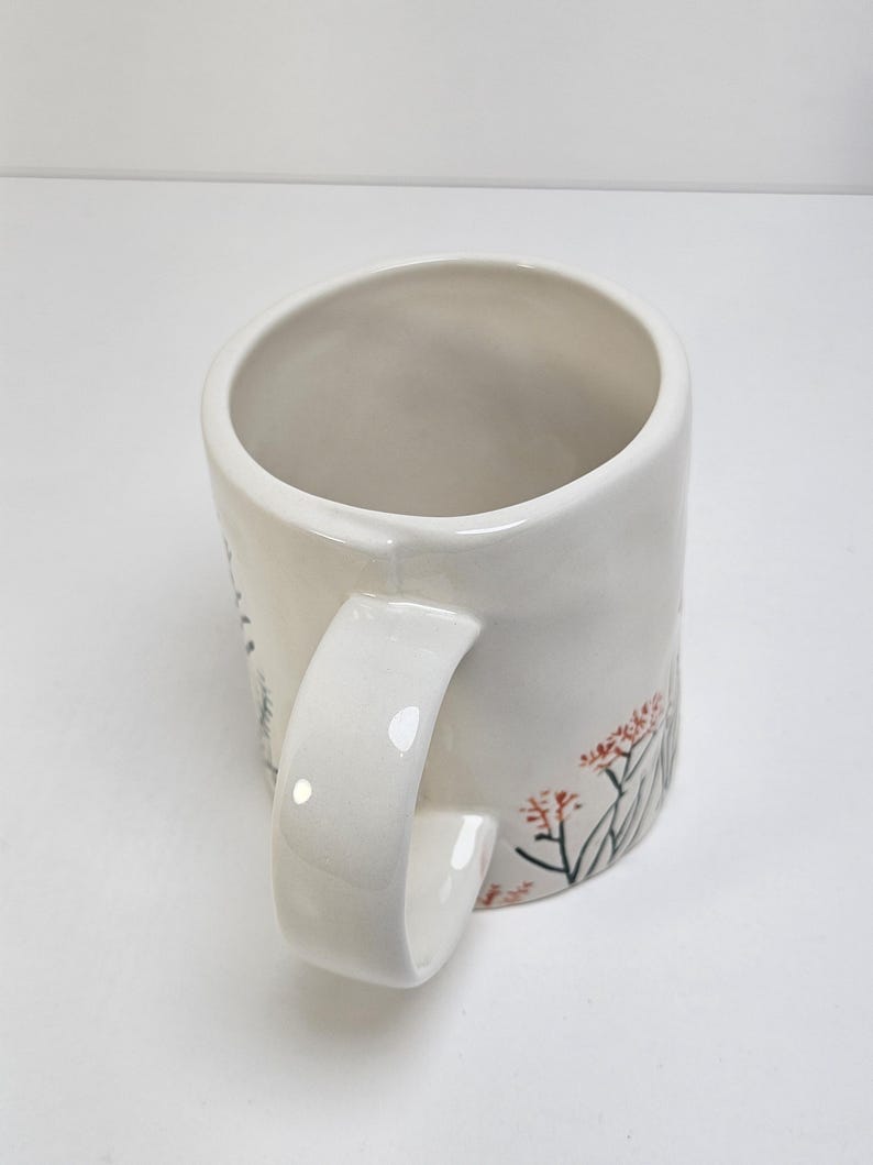 K&ouml;nnte beinhalten: Wei&szlig;er Keramikbecher mit gebogenem Henkel. Der Becher hat ein Blumendesign mit roten und gr&uuml;nen Akzenten. Der Becher ist zylindrisch und hat eine glatte, gl&auml;nzende Oberfl&auml;che. Der Griff ist dick und leicht zu greifen.