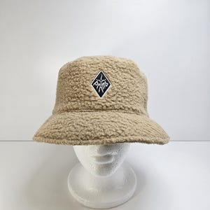 May include: A tan bucket hat with a soft, textured appearance. The hat features a black and white diamond-shaped logo on the front. The hat is displayed on a white mannequin head against a white background.