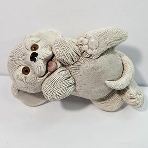 May include: A white ceramic figurine of a puppy lying on its back with its paws in the air. The puppy has brown eyes and a black nose.