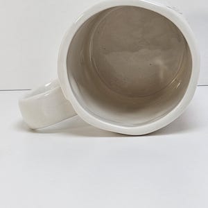 K&ouml;nnte beinhalten: Eine wei&szlig;e Keramiktasse mit rundem Henkel. Das Innere der Tasse ist hellbeige. Die Tasse steht auf einer wei&szlig;en Oberfl&auml;che.