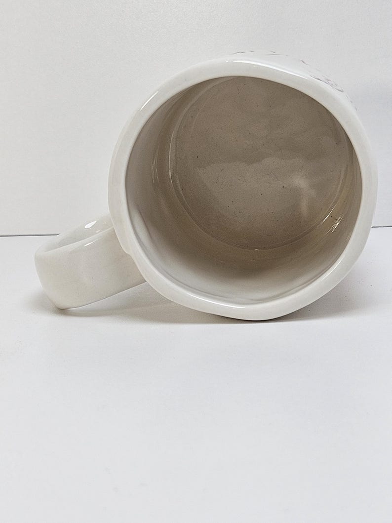 K&ouml;nnte beinhalten: Eine wei&szlig;e Keramiktasse mit rundem Henkel. Das Innere der Tasse ist hellbeige. Die Tasse steht auf einer wei&szlig;en Oberfl&auml;che.