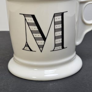 May include: White ceramic mug with a large black letter "M" on the front. The letter is filled with horizontal lines and dashes. The mug has a handle and a pedestal base.