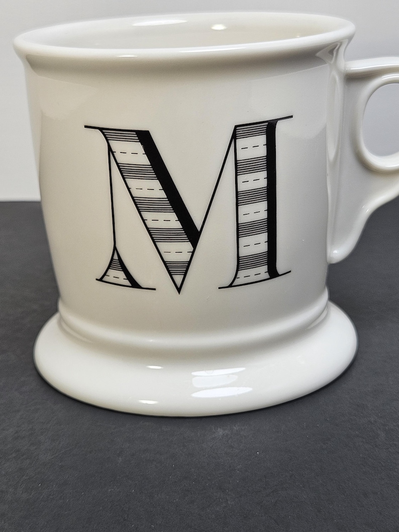 May include: White ceramic mug with a large black letter "M" on the front. The letter is filled with horizontal lines and dashes. The mug has a handle and a pedestal base.