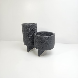 May include: A dual-chambered planter featuring a dark charcoal gray, heavily textured, porous surface. The object consists of two cylindrical vessels of varying heights, each supported by a single, flat, triangular base extending downward.