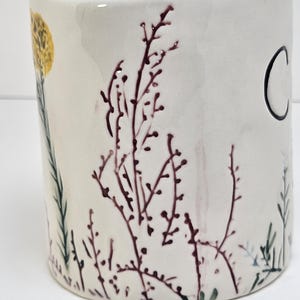 K&ouml;nnte beinhalten: Eine wei&szlig;e Keramiktasse mit zylindrischer Form. Die Tasse ist mit einer gelben Blume, dunkelroten botanischen Illustrationen und dem Buchstaben "C" in Schwarz verziert. Gr&uuml;ne und blaue Pflanzenillustrationen sind ebenfalls vorhanden.