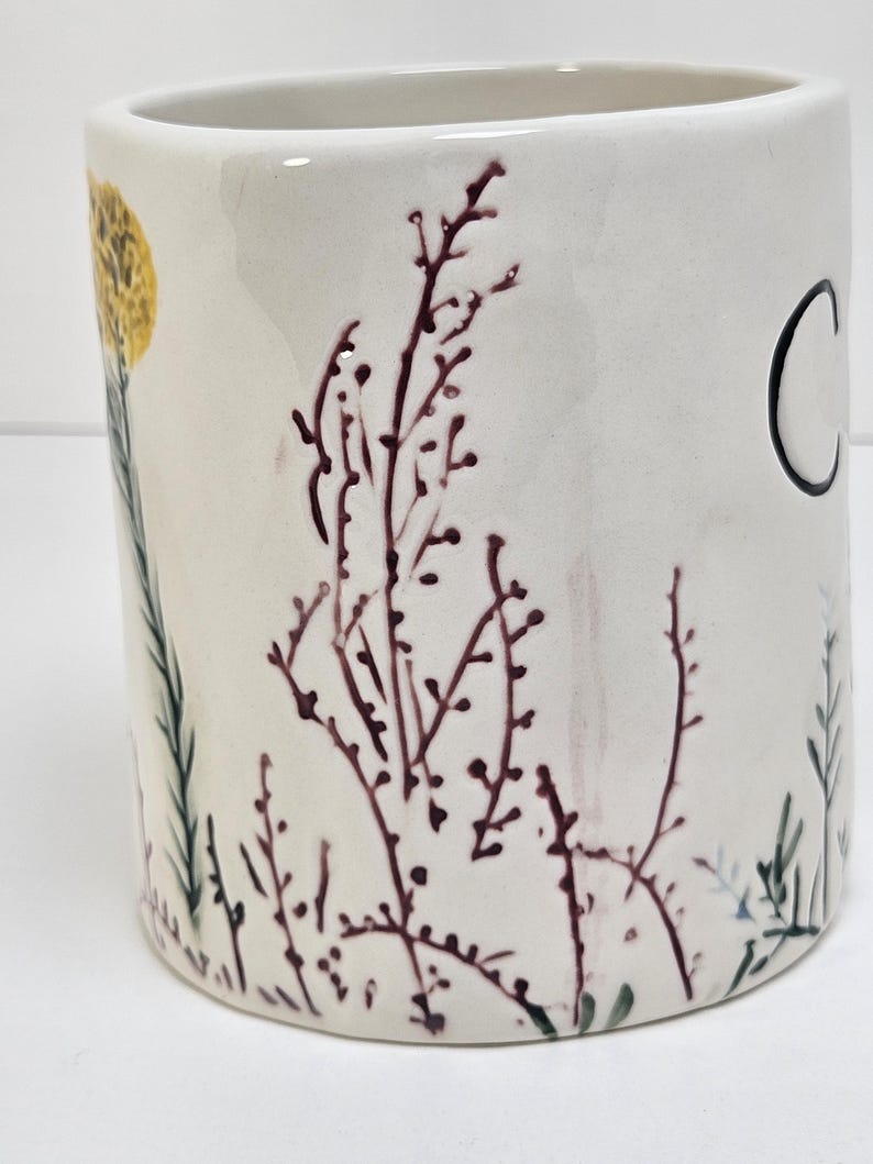 K&ouml;nnte beinhalten: Eine wei&szlig;e Keramiktasse mit zylindrischer Form. Die Tasse ist mit einer gelben Blume, dunkelroten botanischen Illustrationen und dem Buchstaben "C" in Schwarz verziert. Gr&uuml;ne und blaue Pflanzenillustrationen sind ebenfalls vorhanden.