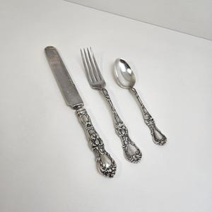 May include: A three-piece silver-toned flatware set consisting of a dinner knife, a four-tine fork, and a spoon. Each piece features an ornate, embossed floral and scrollwork pattern on the handle, contrasting with the smooth, polished metal blades and bowls.