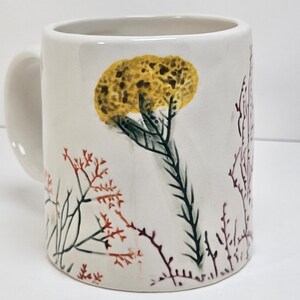 K&ouml;nnte beinhalten: Eine wei&szlig;e Keramiktasse mit gebogenem Henkel. Die Tasse hat ein handgemaltes Design aus gelben und gr&uuml;nen floralen Elementen sowie roten und braunen Zweigen. Die Tasse hat eine Standardgr&ouml;&szlig;e f&uuml;r Kaffee oder Tee.