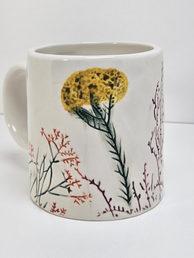 K&ouml;nnte beinhalten: Eine wei&szlig;e Keramiktasse mit gebogenem Henkel. Die Tasse hat ein handgemaltes Design aus gelben und gr&uuml;nen floralen Elementen sowie roten und braunen Zweigen. Die Tasse hat eine Standardgr&ouml;&szlig;e f&uuml;r Kaffee oder Tee.