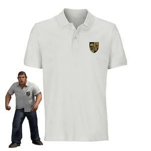 Può includere: Polo bianco con uno stemma blu scuro e oro che presenta un pugno, un teschio, un uccello e uno scudo. Lo stemma è ricamato sul petto sinistro della polo.