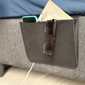 May include: A dark gray bedside storage pocket made of a smooth material, featuring a vertical stitched seam. The pocket holds a black smartphone, a white charging cable, a beige notebook, and a pair of dark-framed glasses resting on the front.