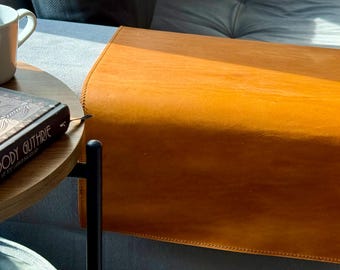 May include: A tan leather armrest tray with stitched edges draped over a gray sofa. Next to it, a round wooden side table holds a white ceramic mug and a book with the text Woody Guthrie visible on the black spine cover.