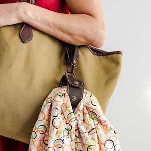 May include: A tan canvas tote bag with brown leather accents and a matching leather scarf holder. The scarf is white with a colorful pattern of orange, green, and black overlapping circles.