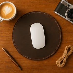 Könnte beinhalten: Ein braunes Leder-Mauspad mit einer weißen Maus, einer Kaffeetasse mit Latte Art, einem Stift, einer Kamera und einem Bindfadenbündel auf einer Holzoberfläche. Das Pad ist rund mit genähten Kanten.