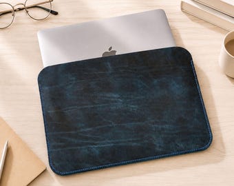May include: A rectangular laptop sleeve featuring a dark blue, distressed, textured finish with visible stitching along the edges. The sleeve is partially holding a silver laptop with a visible apple logo, showcasing a sleek and functional design.