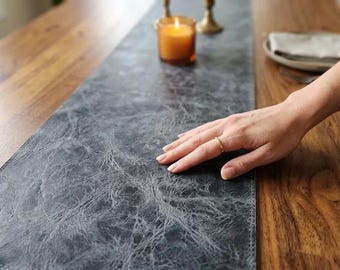 May include: A dark gray faux leather table runner with a textured surface. The runner is placed on a wooden table. A lit candle and a plate with a napkin are in the background.