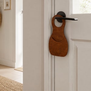 Puede incluir: Un organizador de puerta de cuero marrón con un bolsillo y una abertura para el asa, colgado de una puerta blanca con un tirador negro. El organizador está diseñado para guardar objetos pequeños y es un accesorio decorativo.