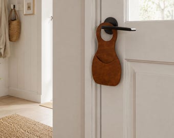 May include: A brown leather door organizer with a pocket and a handle opening, hanging on a white door with a black handle. The organizer is designed to hold small items and is a stylish home decor accessory.