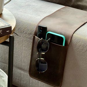 May include: A brown leather couch caddy with a pocket holding a black remote, a teal-edged smartphone, and sunglasses. The caddy is draped over the arm of a gray couch. A book and a mug are on a side table.