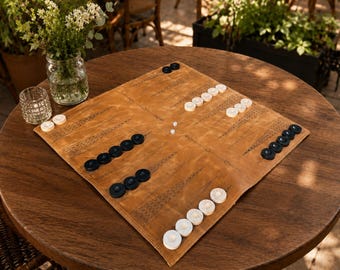 May include: A square, tan leather backgammon board featuring dark brown printed triangular points. The board is set with a collection of circular black and white playing pieces arranged in various stacks across the surface, along with two small white dice.