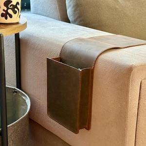 May include: A brown leather sofa armrest organiser with a pocket. The organiser is designed to fit over the arm of a sofa. The leather is a rich brown colour, and the pocket is rectangular.