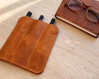 May include: A leather pen case with three slots, holding three black pens. A brown leather notebook with glasses rests on a wooden surface. The pen case and notebook are a similar shade of brown.