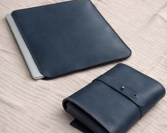 May include: Two navy blue leather cases. One is a flat, rectangular sleeve with a laptop inside. The other is a folded case with a strap and button closure. Both cases are on a light beige surface.