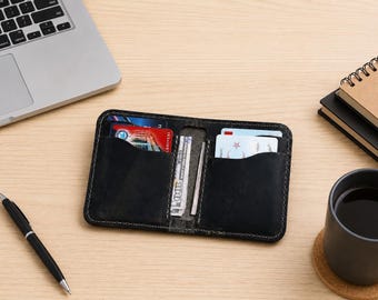 May include: Black leather wallet with multiple card slots and a cash compartment. The wallet is open, revealing credit cards and a wad of US currency. A pen, laptop, notebook, and a cup of coffee are on the wooden surface.
